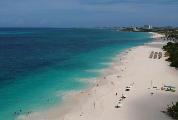 Aerial view of a stunning Caribbean beach with turquoise and blue waters, white sand, palapas, and a distant town under a blue sky.