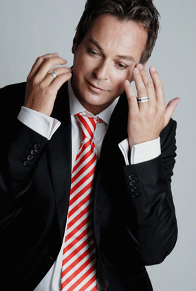 julian clary comedian in black suit red striped tie hands to face